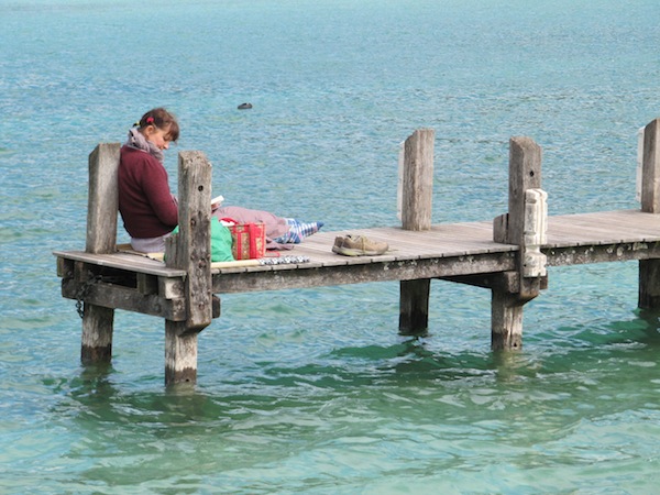 A woman sits enjoying the sun and reading a book on a jetty.