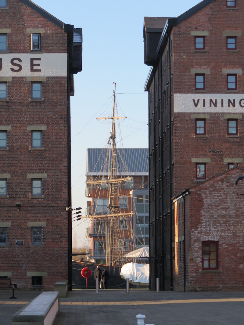 A sail boat is docked visible between two old warehouses